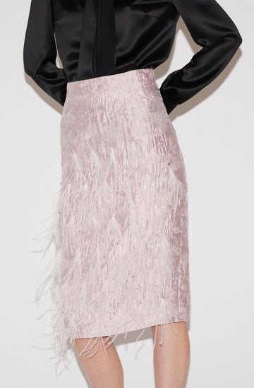 A model wearing a light pink feathered velvet pencil skirt with a black satin long-sleeve top, shown from the chest down against a plain white background.