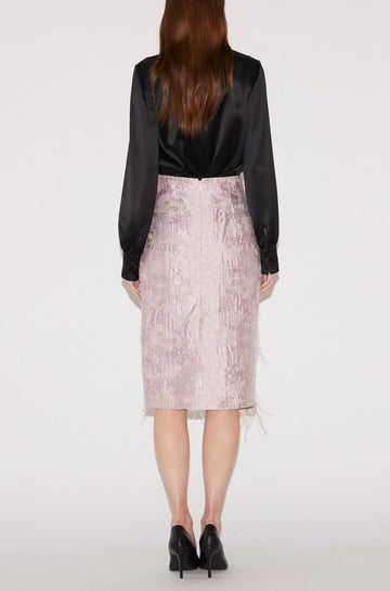 A model viewed from behind wearing a black satin long-sleeve top and a textured light pink skirt with a tulle hem, paired with black heels, against a plain white studio background.
